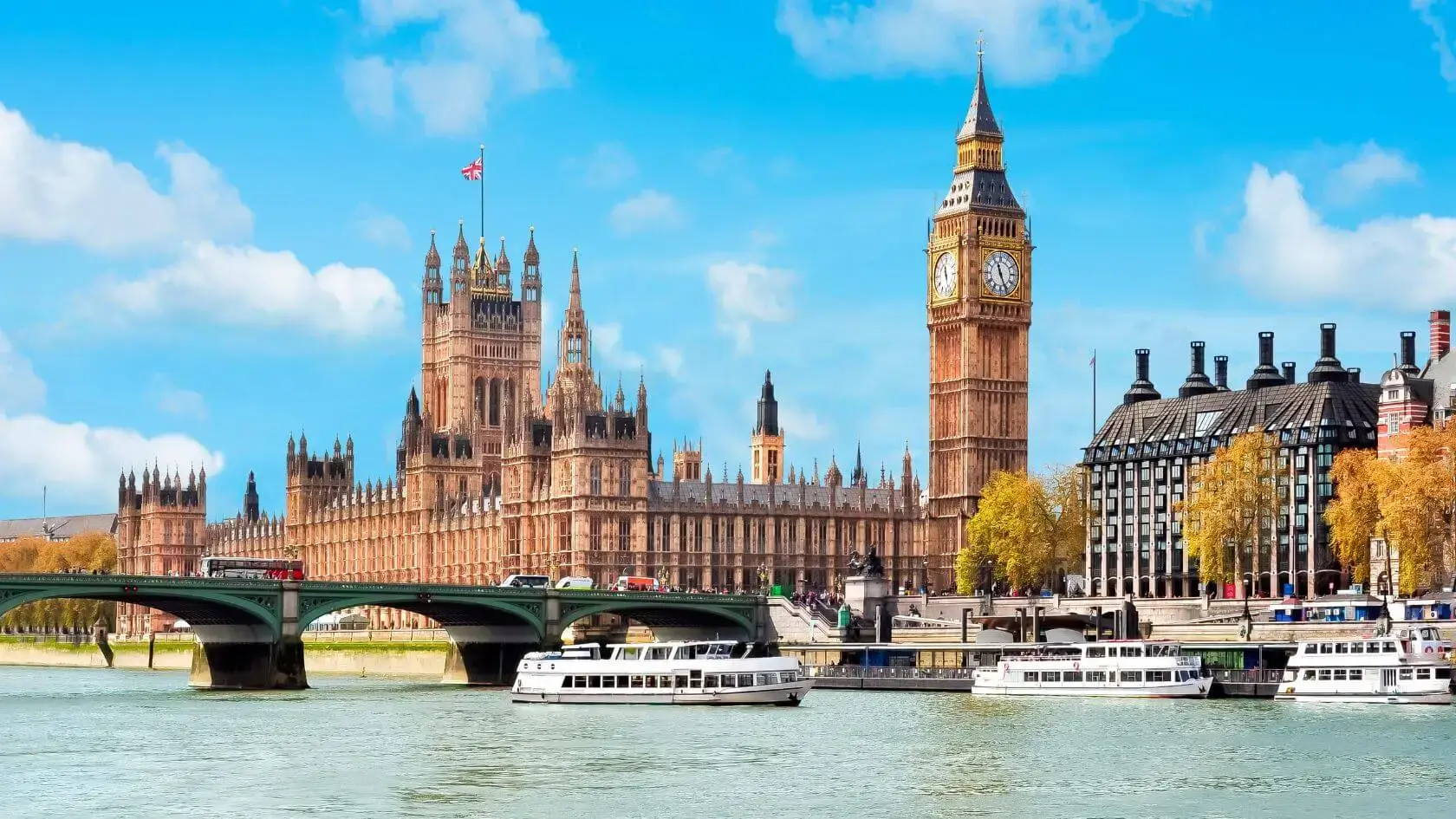 A scenic view of the Houses of Parliament and Big Ben in London, UK, with the Westminster Bridge crossing the River Thames. Boats are seen on the water, and the sky is bright blue with scattered clouds.